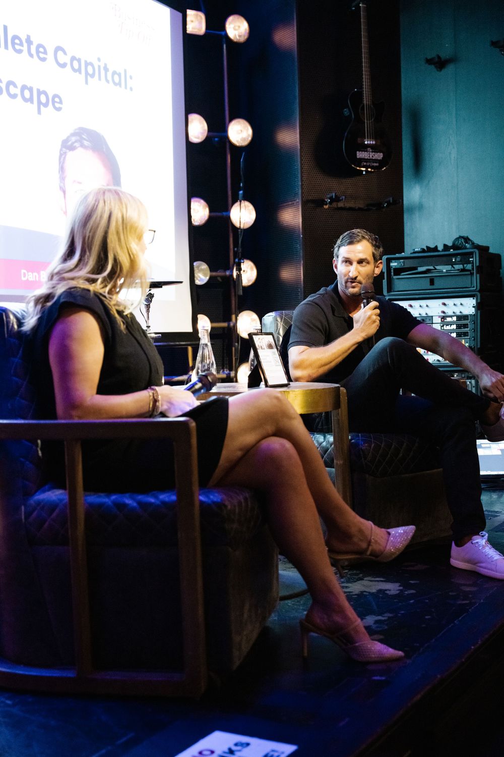 Two people sit in conversation on a dimly lit stage with illuminated vanity lights and audio equipment visible in background.