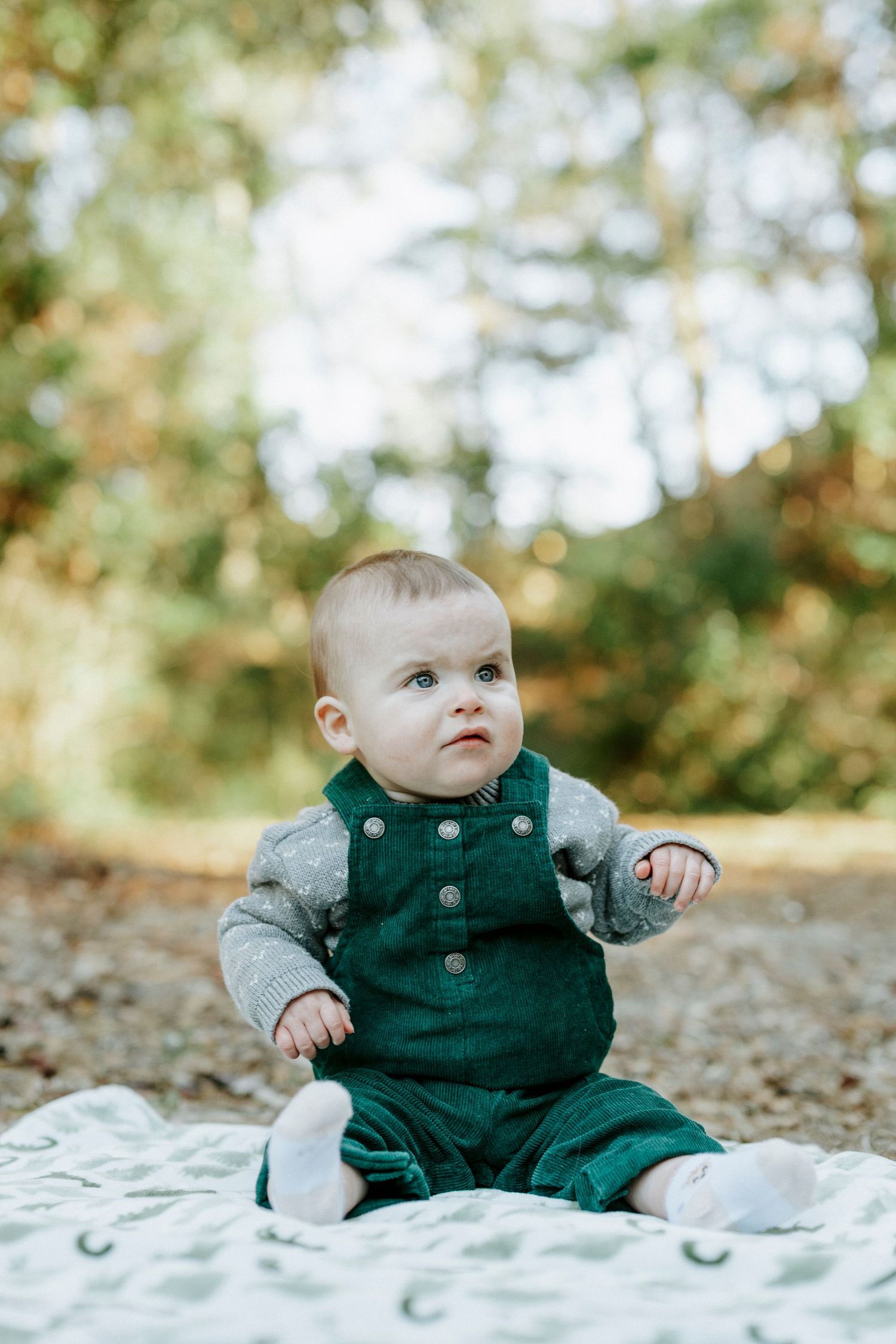 Baby in green overalls sits on white blanket outdoors during autumn photoshoot.