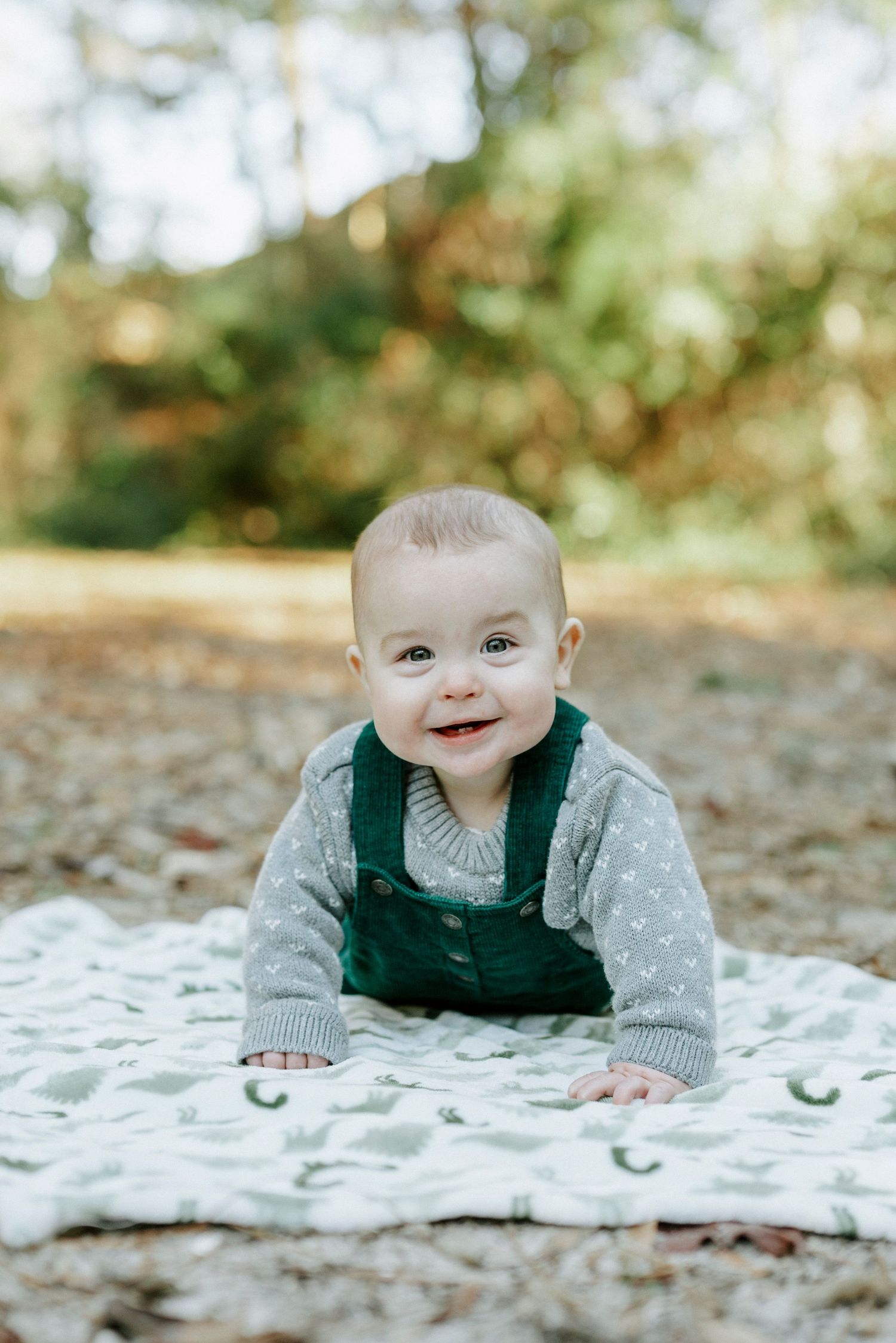 A baby in a green vest and gray sweater lies on a white blanket outdoors during an autumn photoshoot.