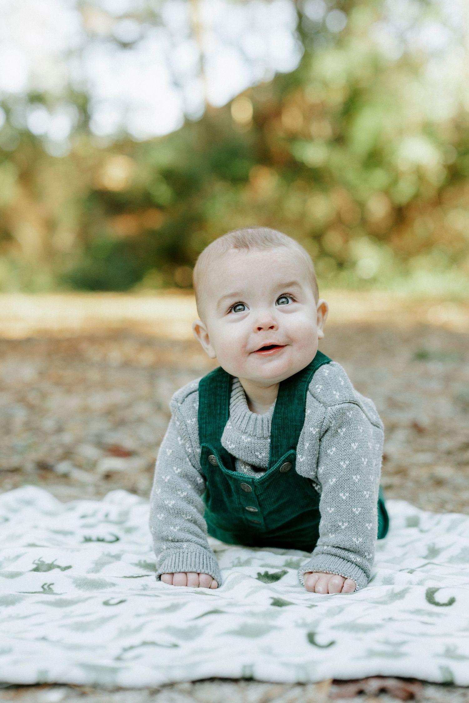 Baby wearing green vest and gray sweater lying on patterned blanket in outdoor setting.