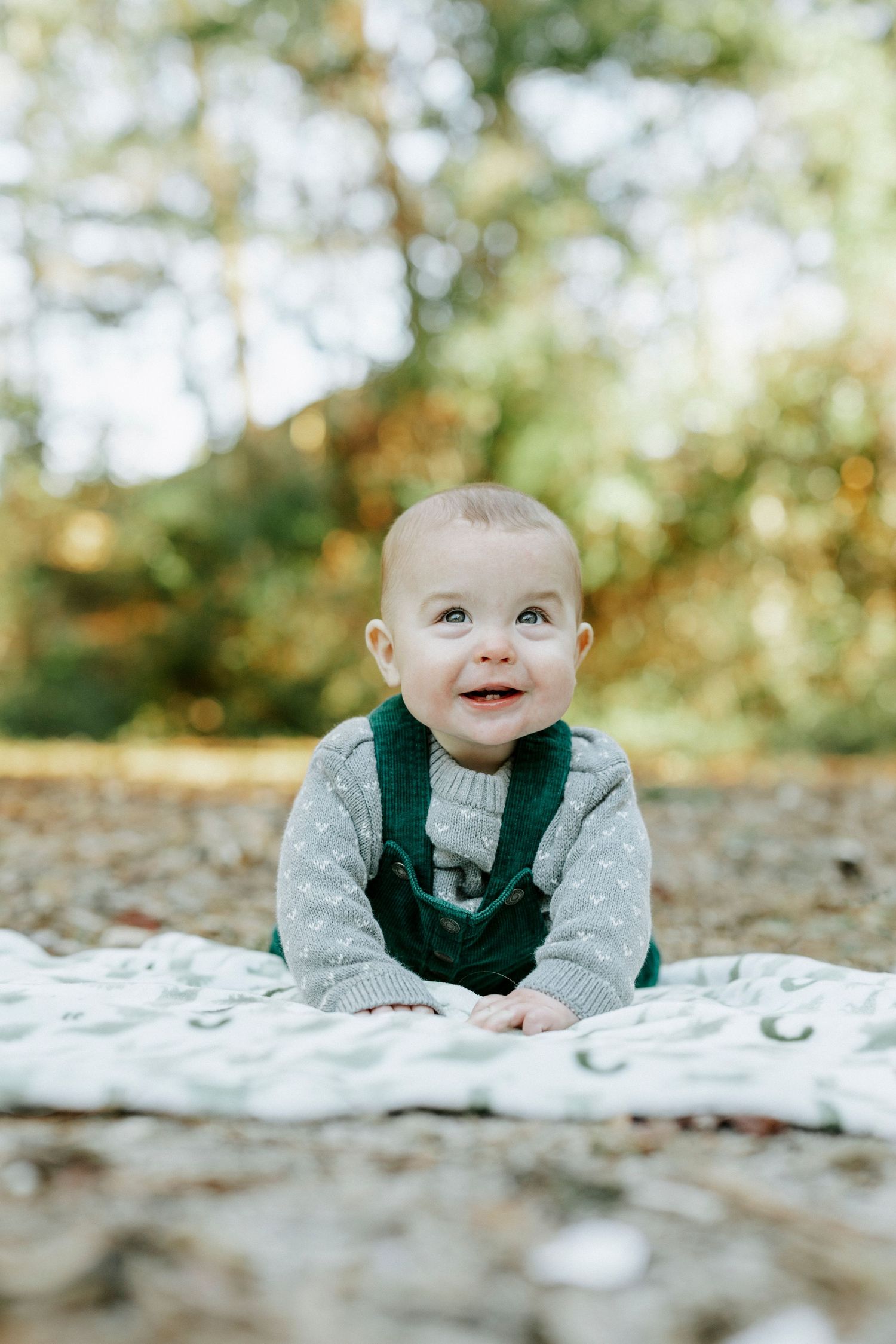 Baby lying on white blanket outdoors smiling with autumn foliage in background.