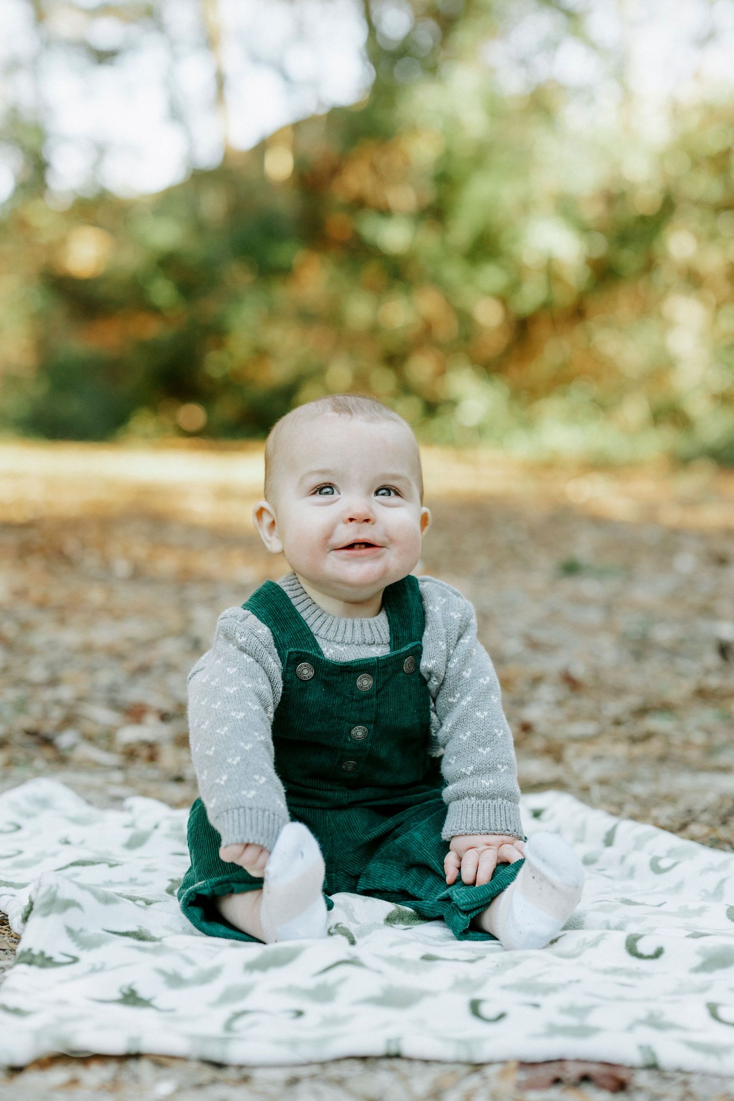 A baby in a dark green overalls and gray sweater sits on a white blanket surrounded by autumn leaves.