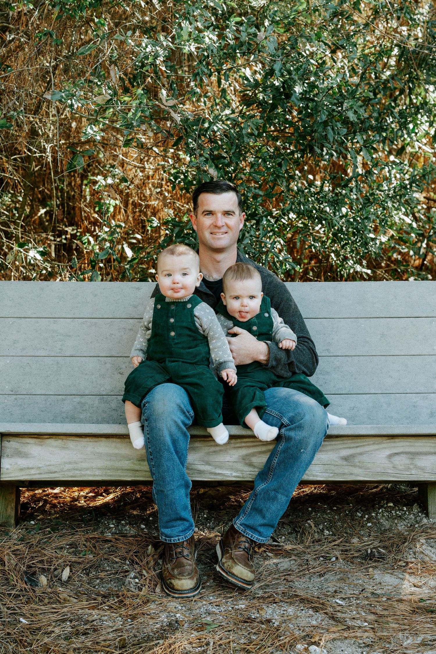 Family sits together on wooden bench surrounded by green foliage during outdoor portrait session.