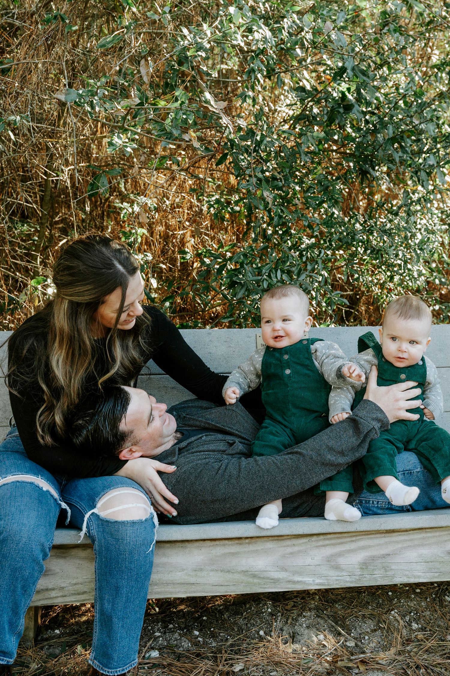 Family relaxing together on wooden bench surrounded by green foliage.