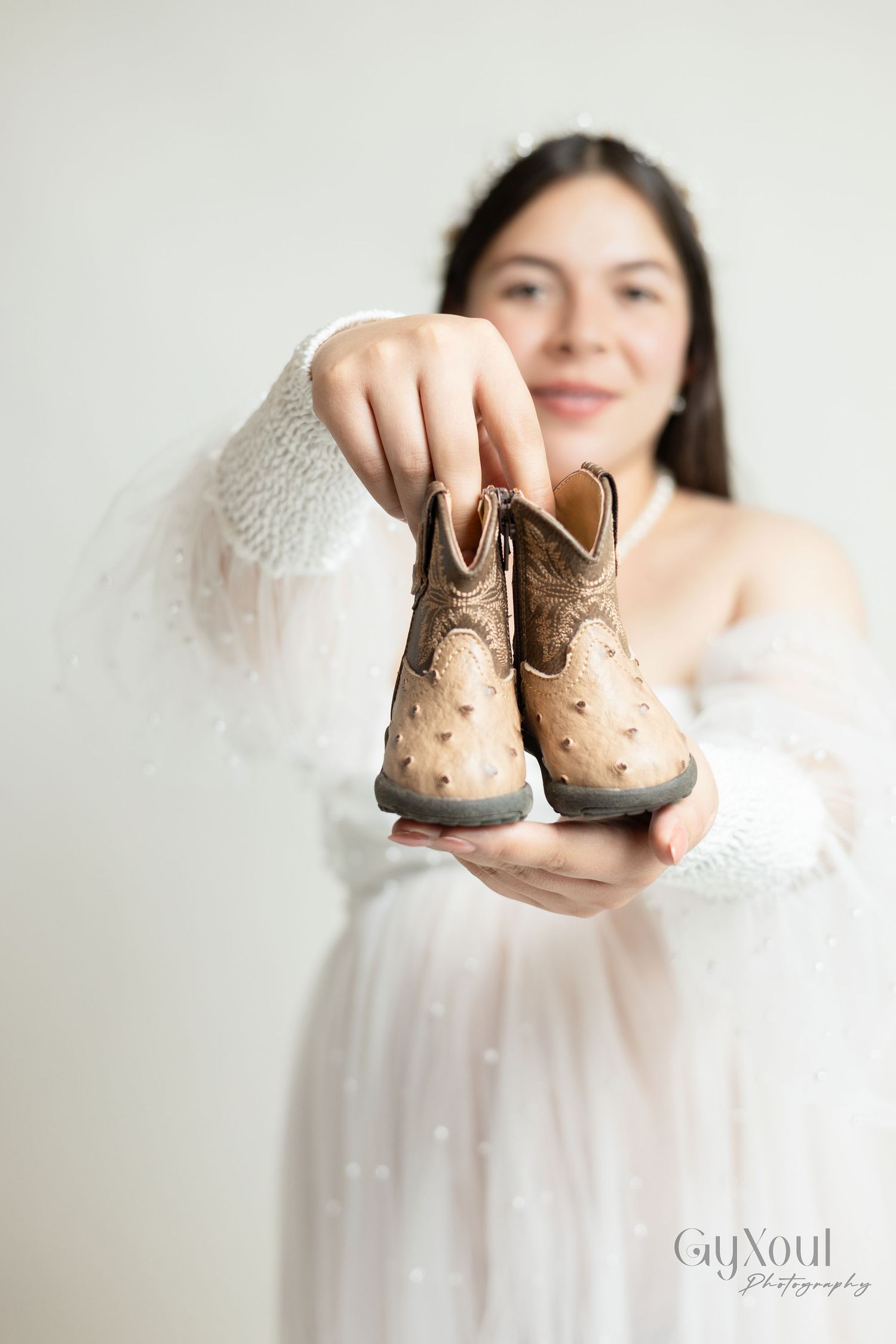 Person in white dress holding tiny decorative cowboy boots against light background.
