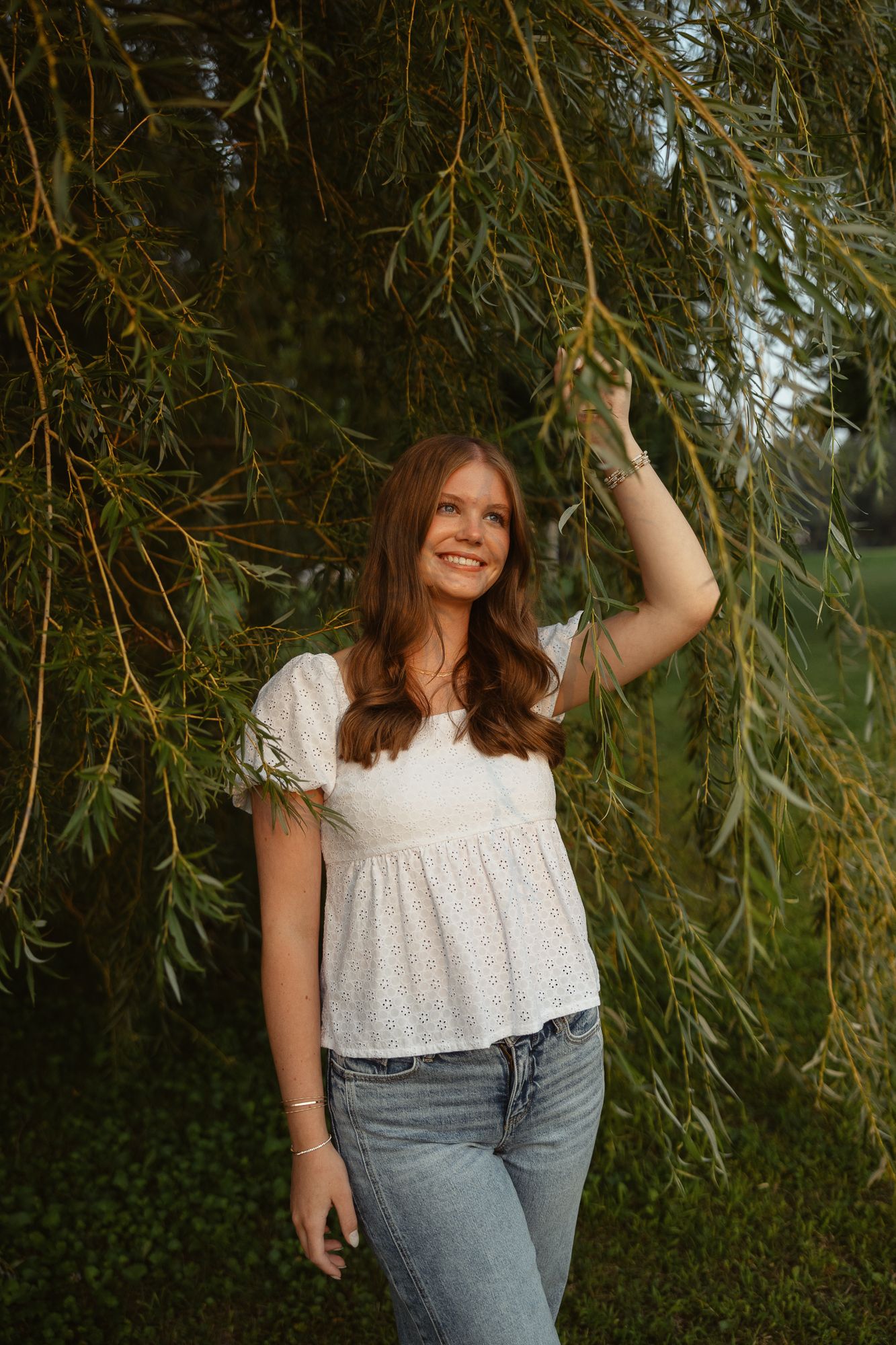 A woman in a white eyelet top and jeans poses playfully under willow tree branches in an outdoor setting.