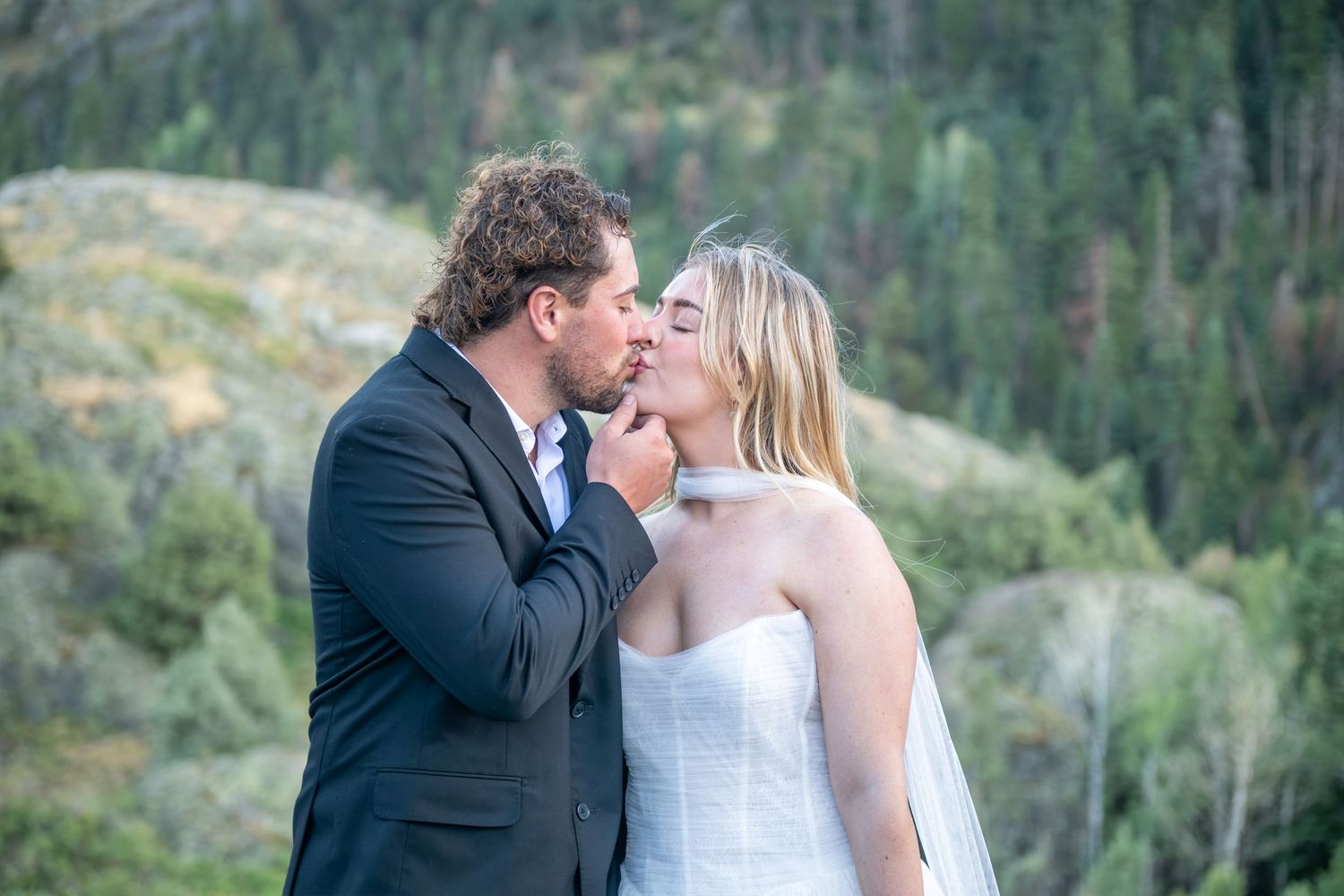An eloping couple in wedding attire sit together in a grassy meadow and share a champagne toast surrounded by golden aspen trees.