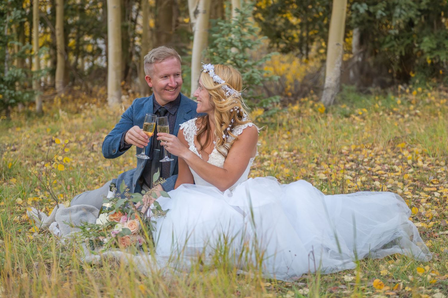 An eloping couple in wedding attire sit together in a grassy meadow and share a champagne toast surrounded by golden aspen trees.