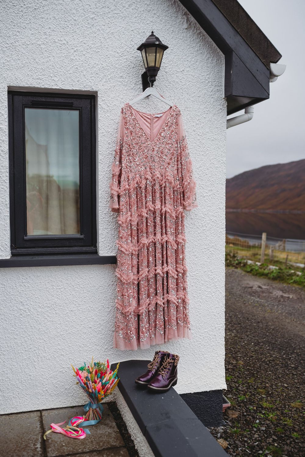 A pink floral dress hangs outside a white cottage with mountains and rugged landscape in the background.