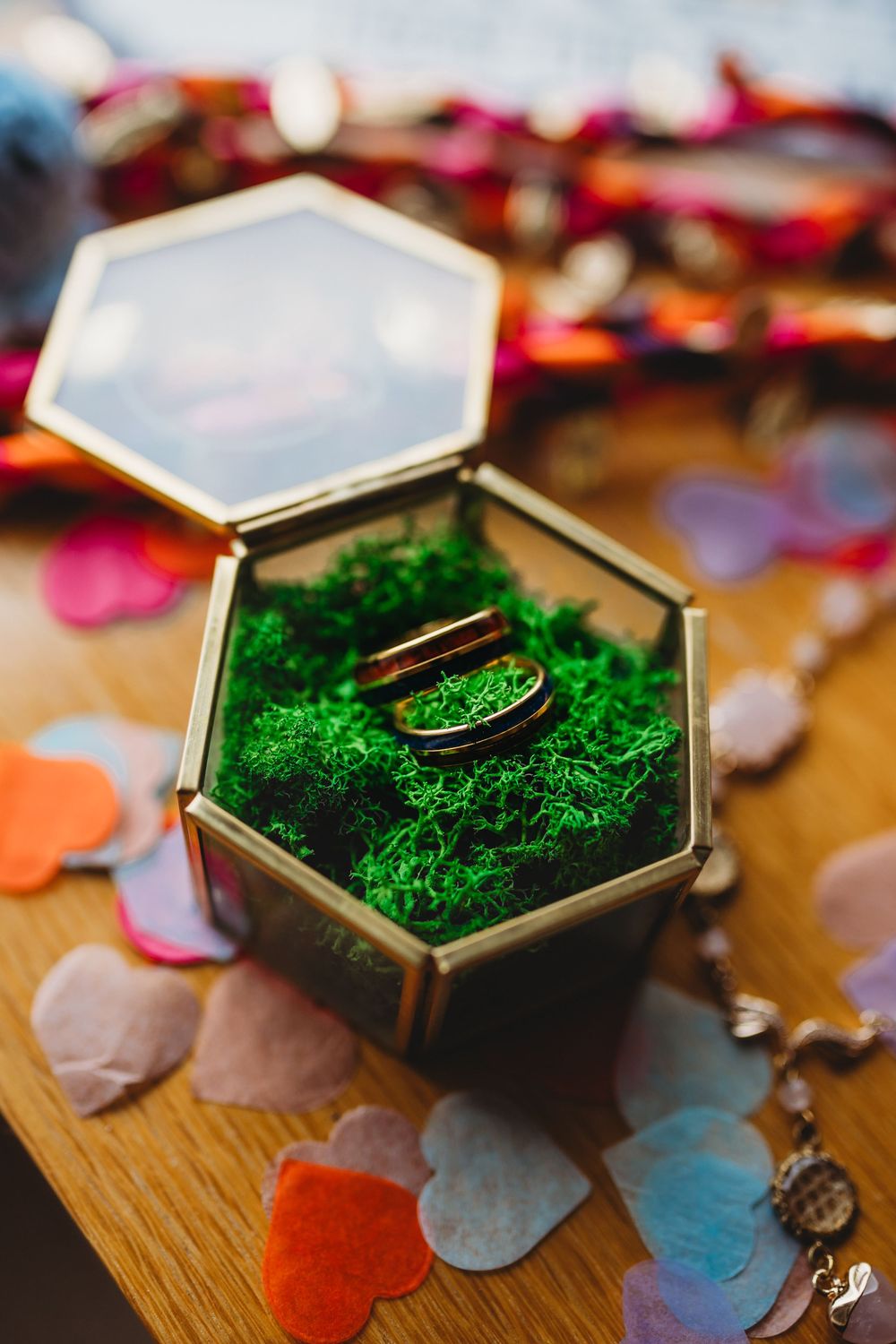 A series of hexagonal boxes with green moss and rings displayed on a wooden surface with scattered paper hearts.