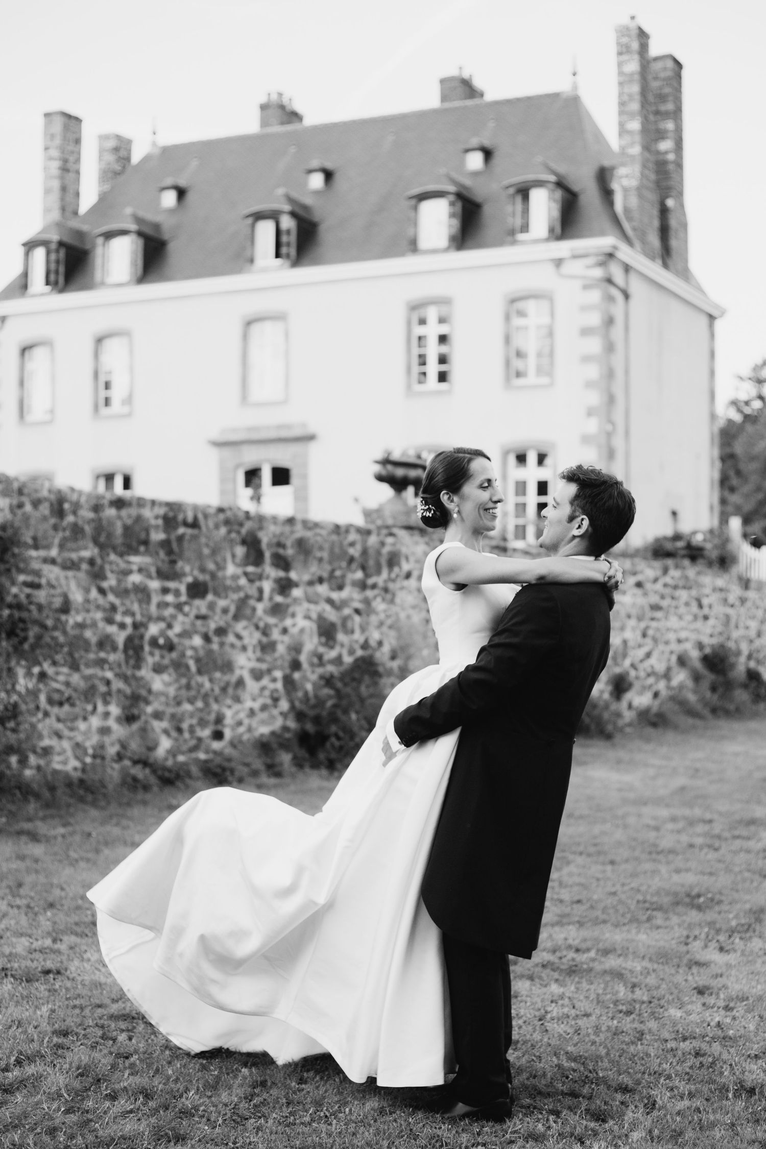 Black and white photo of groom lifting bride in white gown outside elegant French chateau.