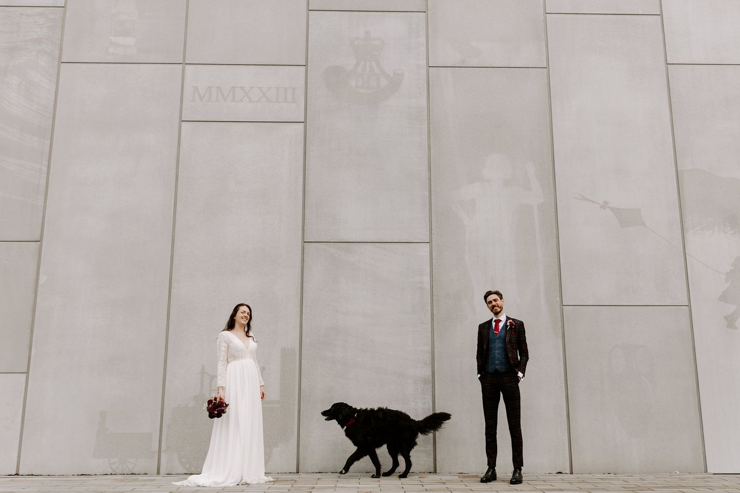 A black dog walks alongside a couple in formal wedding attire against a modern stone wall backdrop.