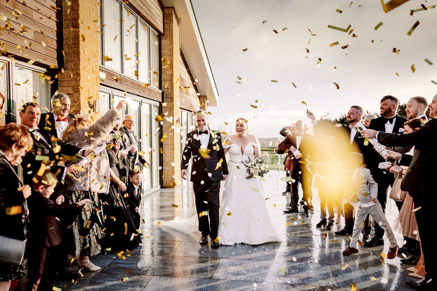 Wedding guests toss glittering confetti as a joyful couple walks down a sunlit hallway lined with cheering onlookers.