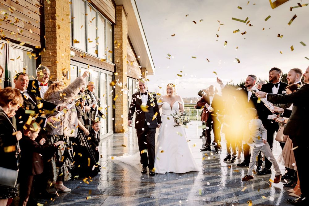 Wedding guests toss glittering confetti as a joyful couple walks down a sunlit hallway lined with cheering onlookers.