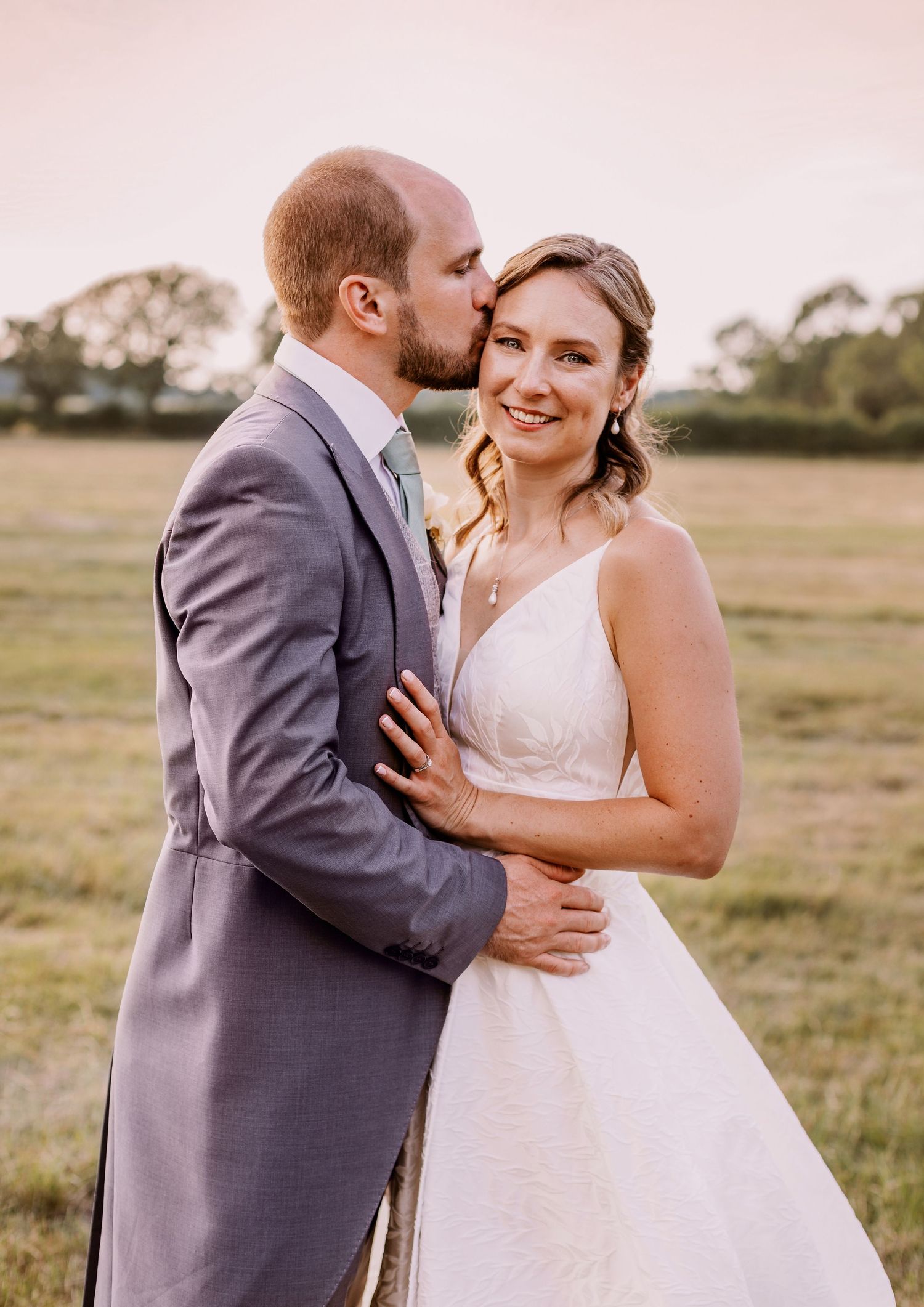 A romantic wedding couple embraces in a field at sunset, with the groom in grey suit and bride in white gown.