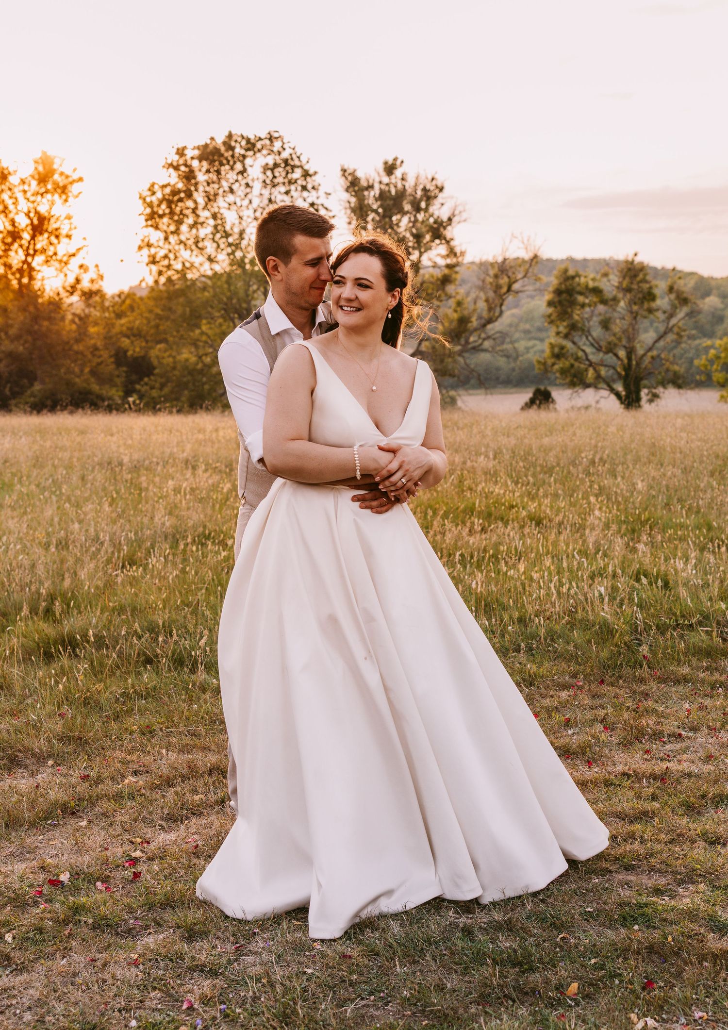 Couple in wedding attire embraces during golden hour in a rustic field at sunset.