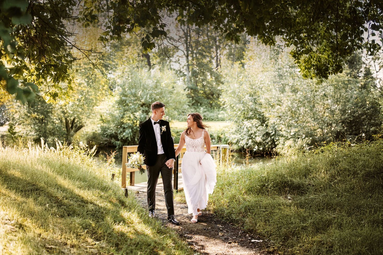 A couple in formal attire walk together on a sunlit woodland path during a wedding photo session.