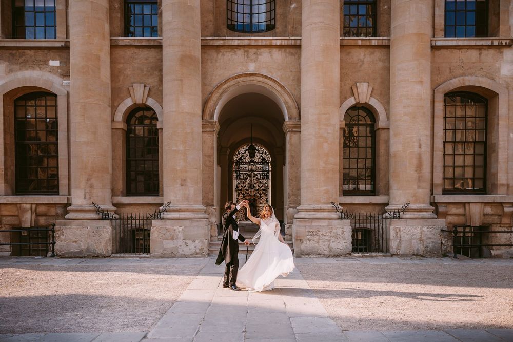 Elegant couple poses in front of ornate stone building with arched windows and classical architecture details.