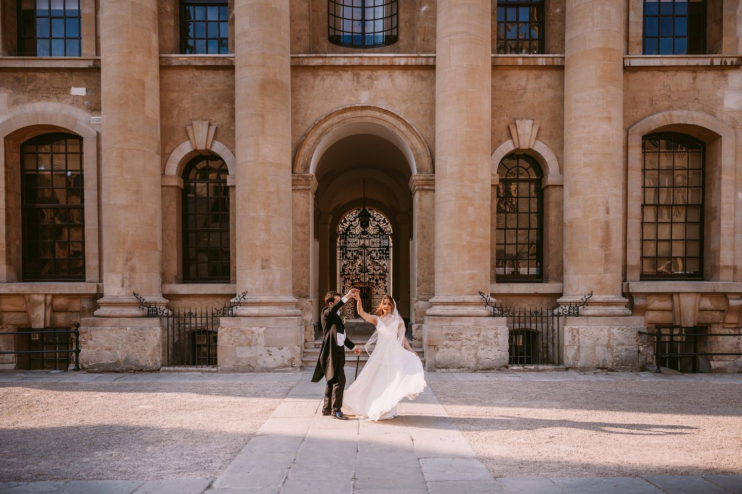 Elegant couple poses in front of ornate stone building with arched windows and classical architecture details.