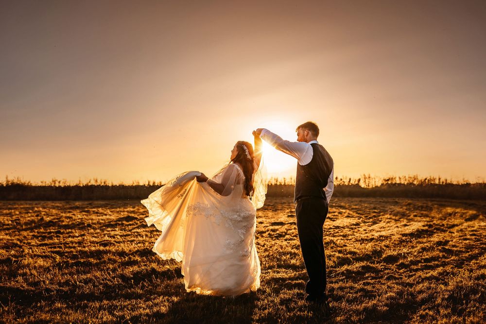Wedding couple poses in dramatic silhouettes across a sun-drenched field at golden hour.