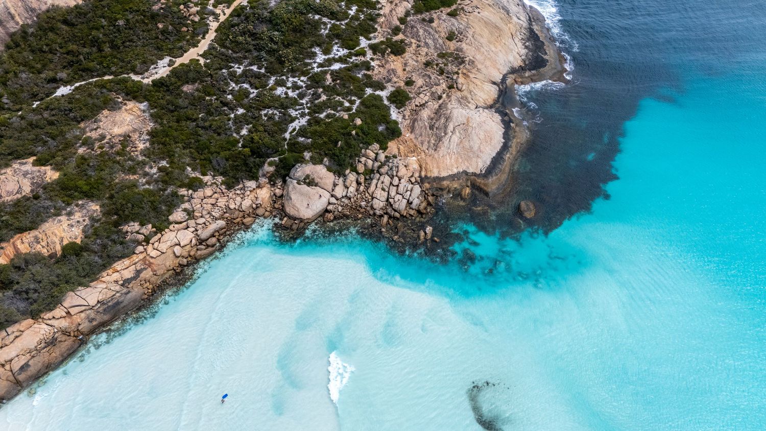Aerial view of crystal clear turquoise water meeting rocky coastline with white sand.