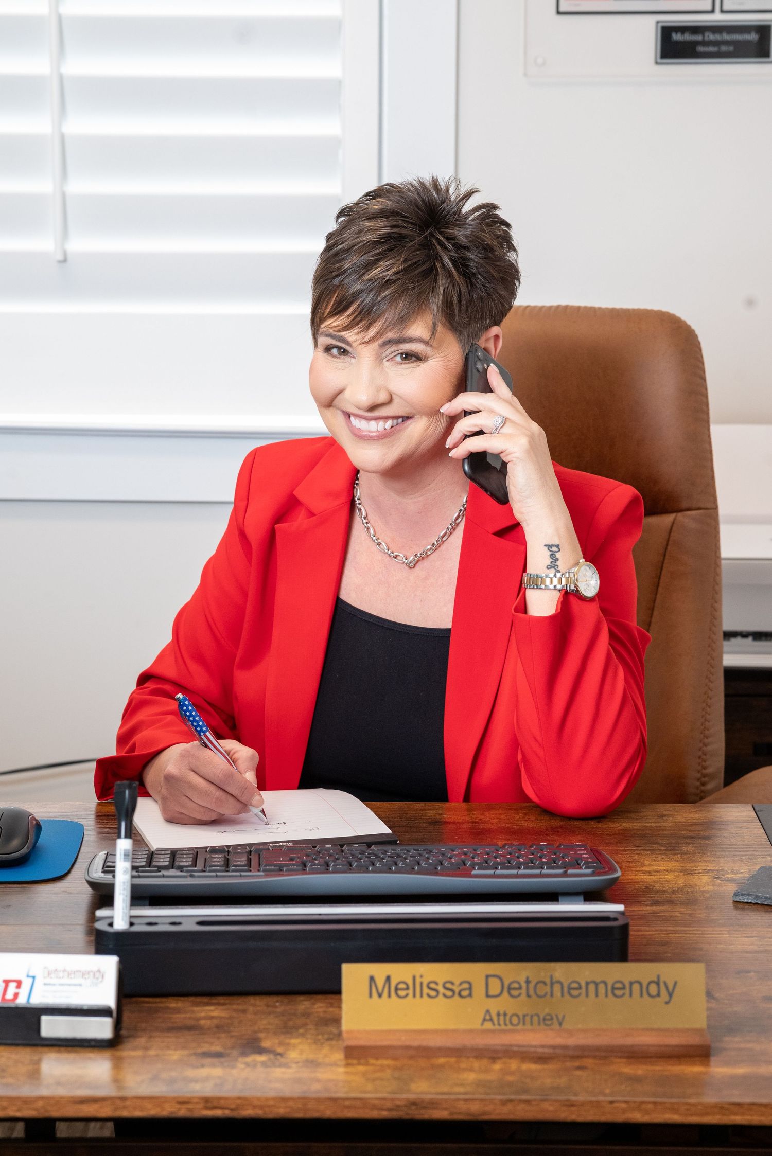 Professional in a red blazer and black top works at wooden desk with nameplate in modern office setting.