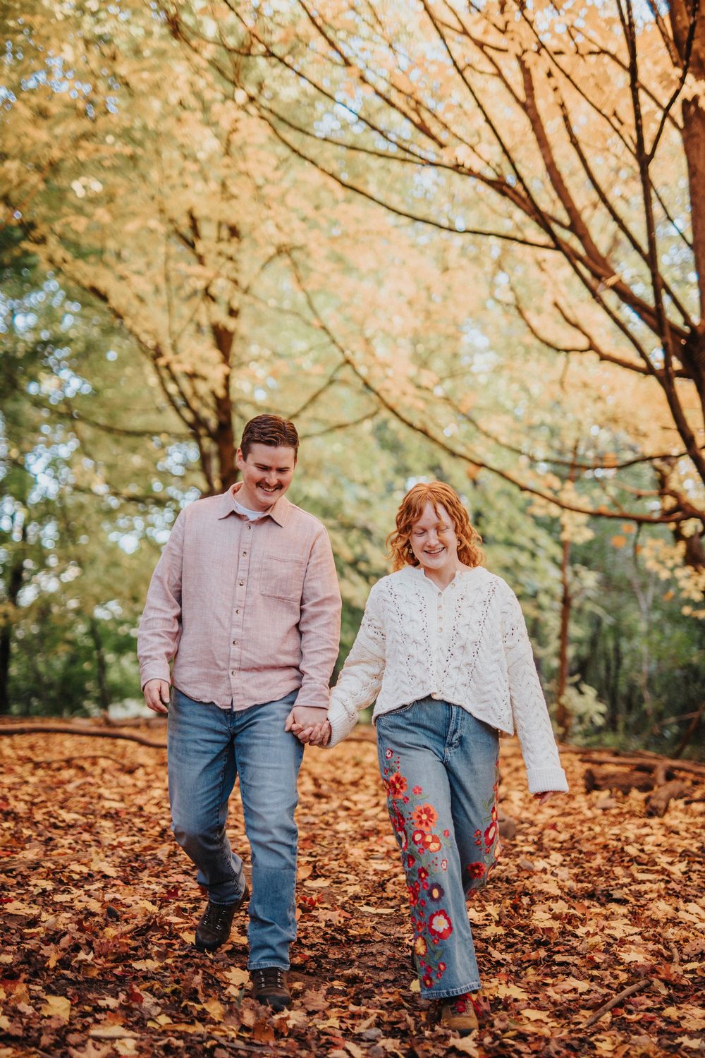 Couple walking through autumn leaves on ground wearing jeans and casual attire in a park setting.