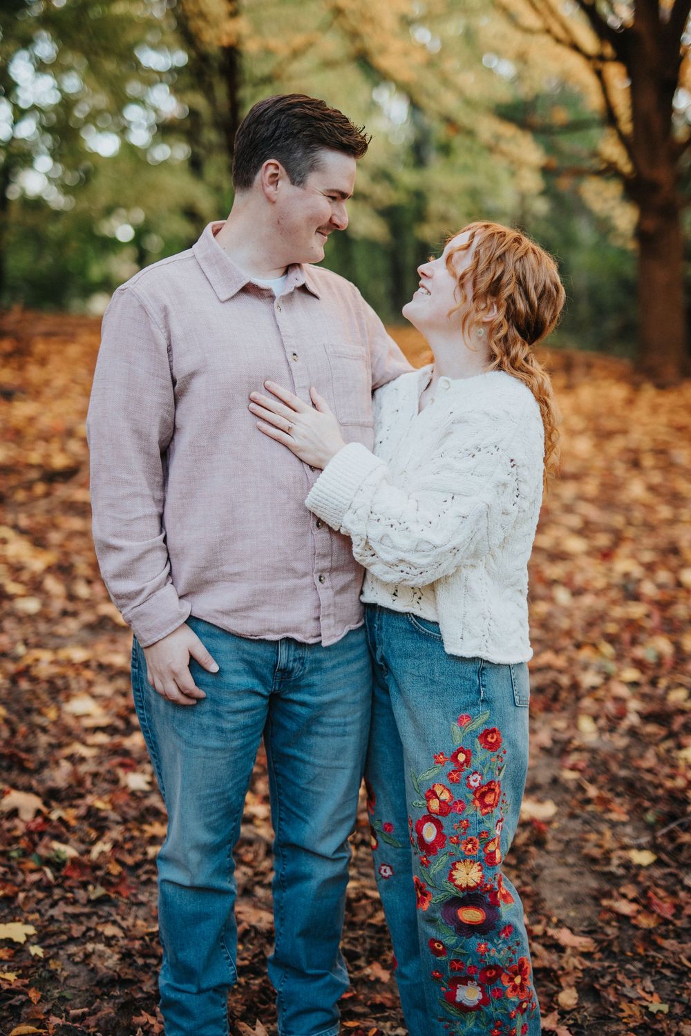 Couple in casual autumn outfits standing among fallen leaves in a park during a romantic photoshoot.