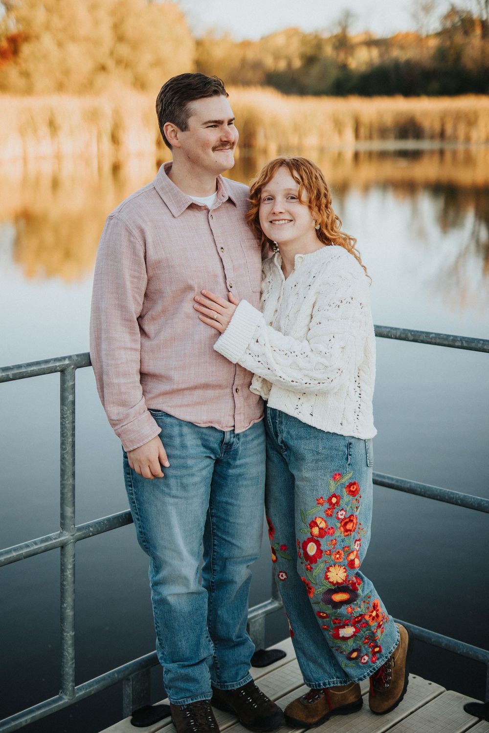 A couple shares a romantic moment on a dock by a lake during golden hour, wearing casual attire and embracing.