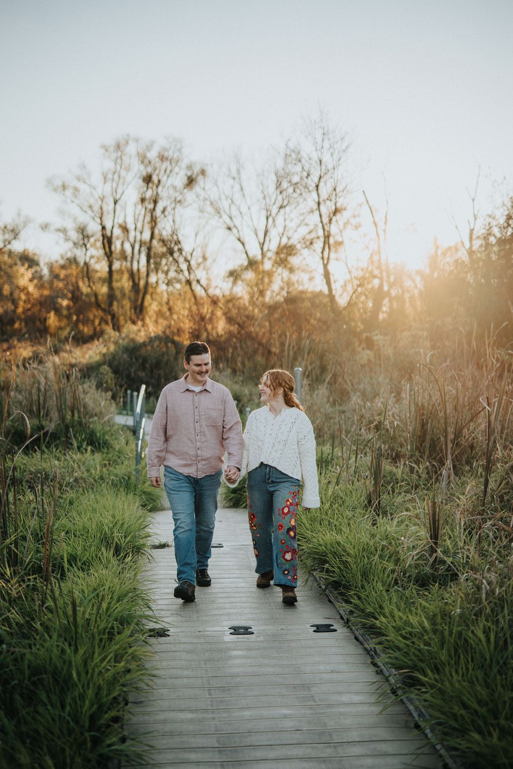 Couple walking together on a wooden boardwalk through tall grass during golden hour sunset.