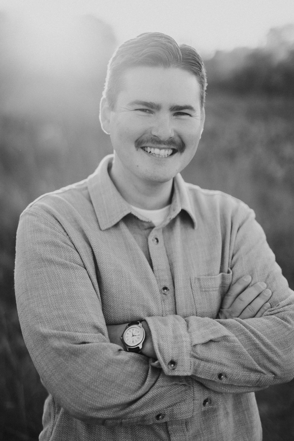 A black and white portrait of a smiling person in a button-down shirt with crossed arms.