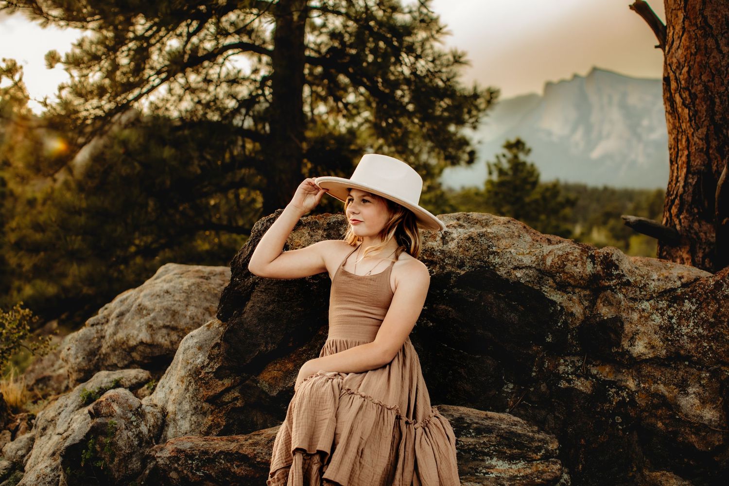 A couple embraces in warm sunset light on a rocky mountain overlook with dramatic mountain peaks in the background.