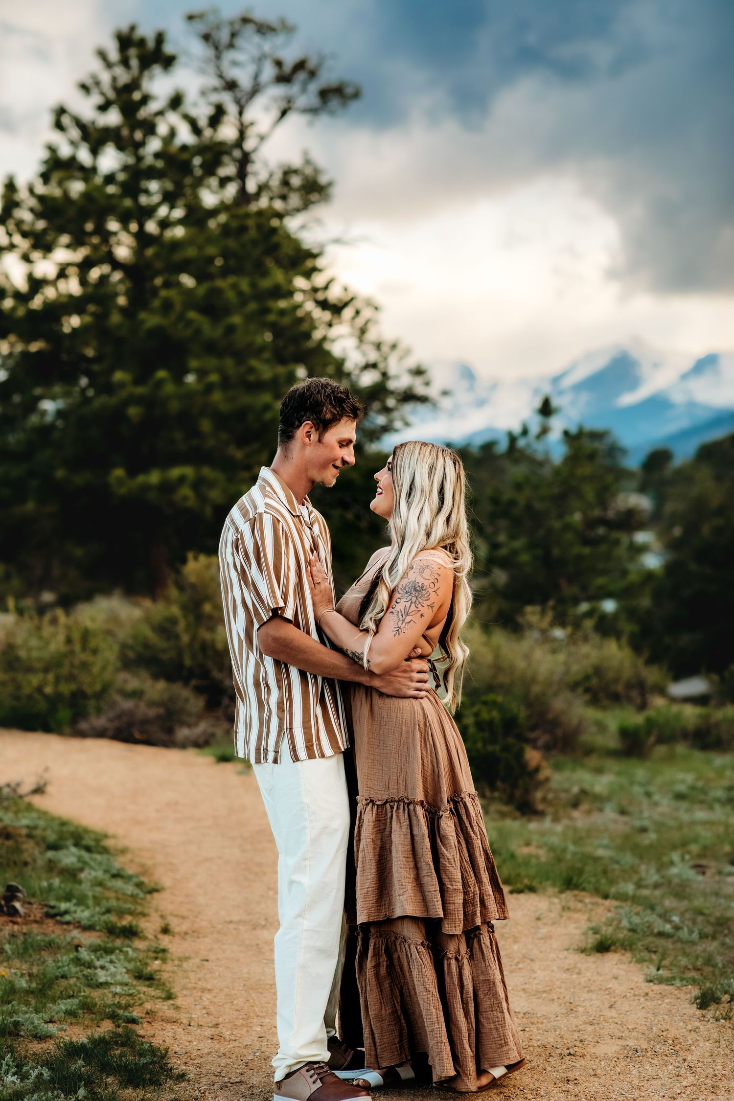 A couple shares a romantic moment on a dirt path at sunset with mountains and trees in the background.
