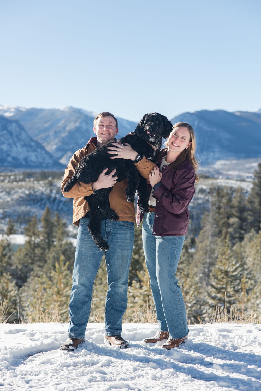 A playful couple poses together in the snow with mountains in the background.