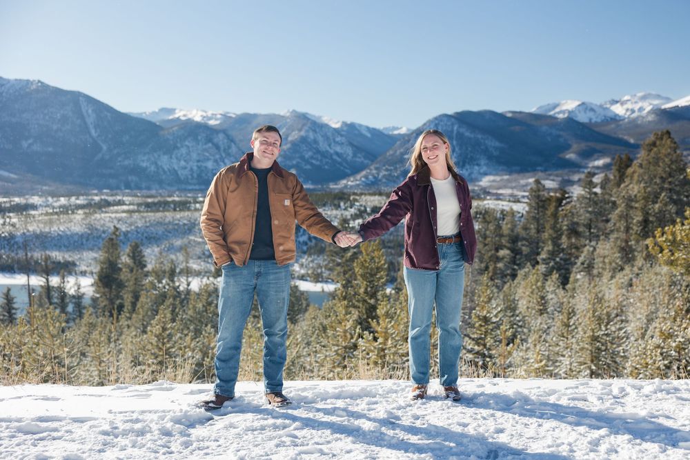 People stand in snow-covered mountain landscape with evergreen trees and scenic winter views in the background.