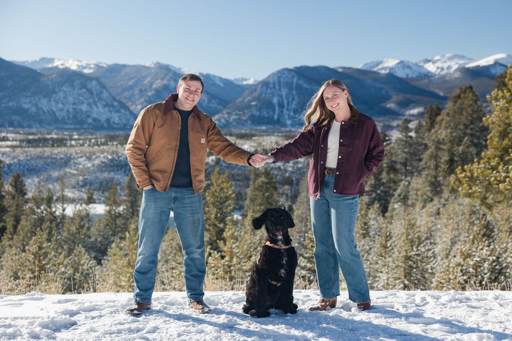 A couple and their black dog pose together in a snowy mountain landscape with evergreen trees and peaks in the distance.