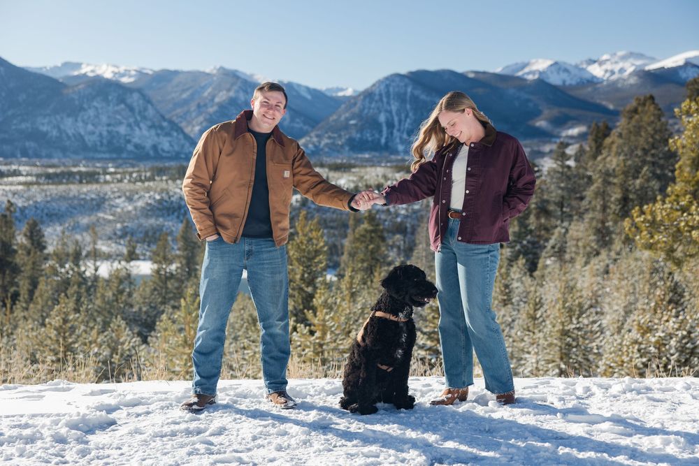 A couple poses together in a snowy mountain landscape with evergreen trees and peaks in the background.