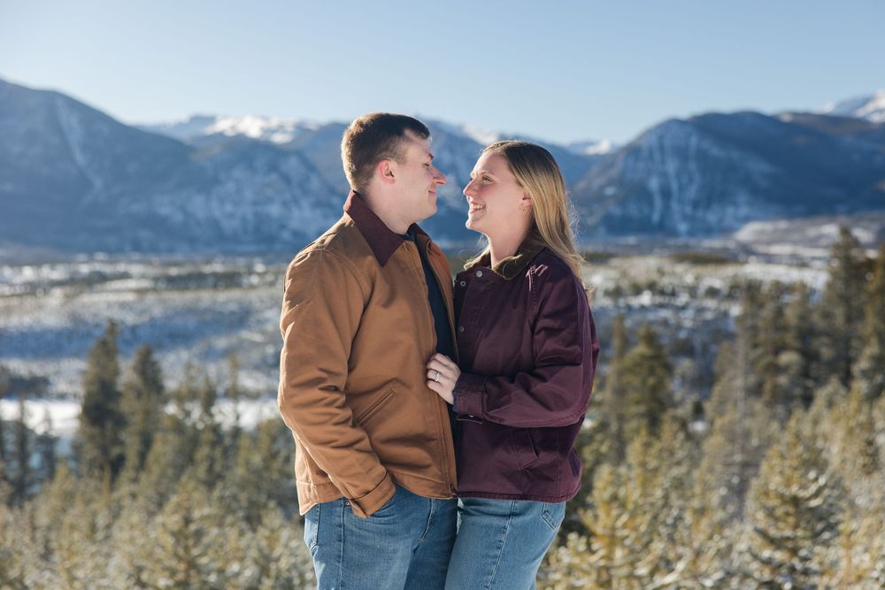 A couple shares an intimate moment against a backdrop of snow-capped mountains and evergreen forest.