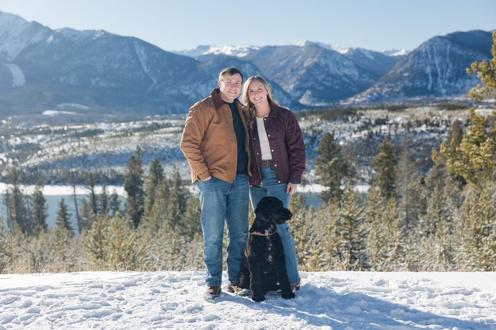 A couple and their black dog pose on a snowy overlook with mountain peaks and a valley town in the background.