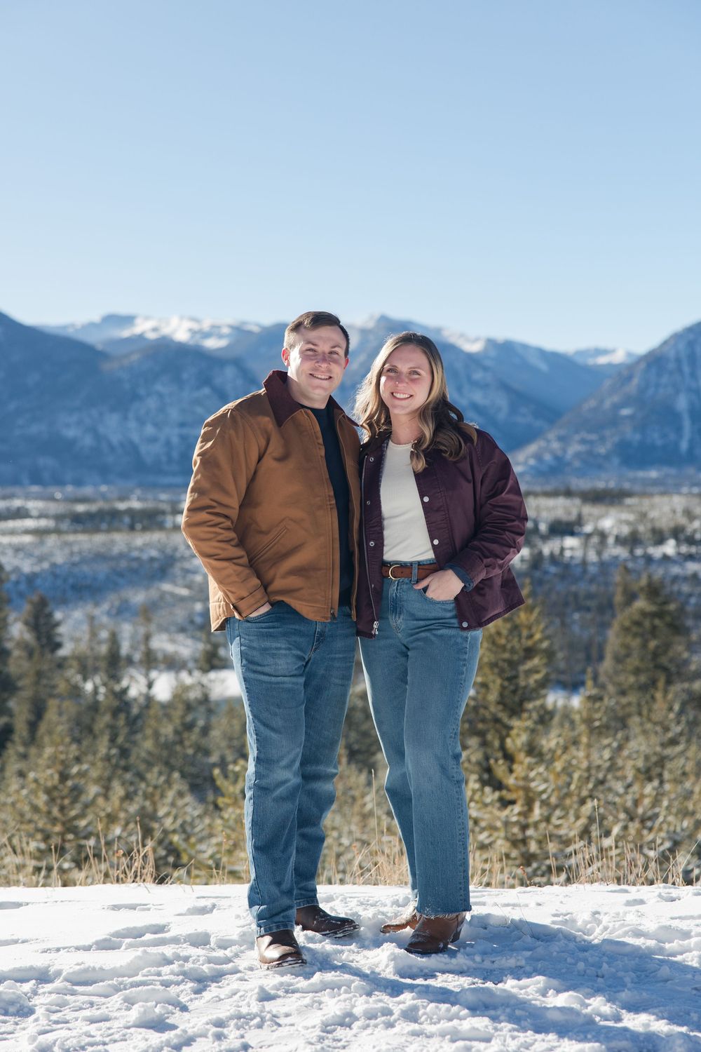 A couple stands together on snowy ground with snow-capped mountains and evergreen forest in the background.