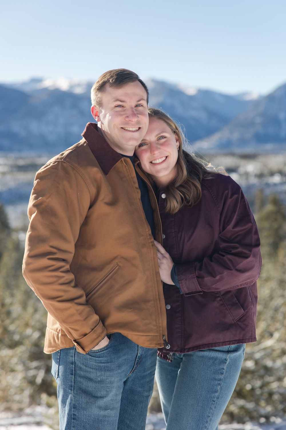 A couple embraces while wearing winter clothing against a snowy mountain backdrop.