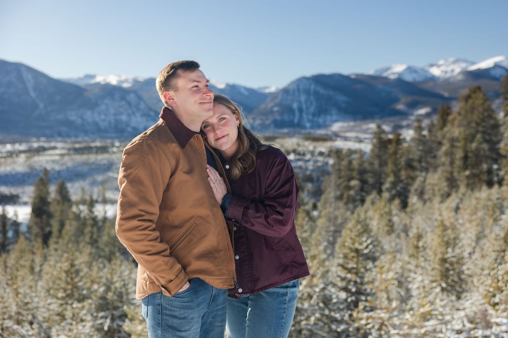 A couple shares an intimate moment against a snowy mountain backdrop with evergreen trees in winter.