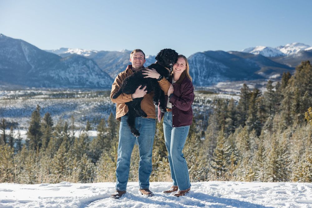 A couple shares a playful moment on a snowy mountain overlook with scenic snow-capped peaks and a lake in the distance.
