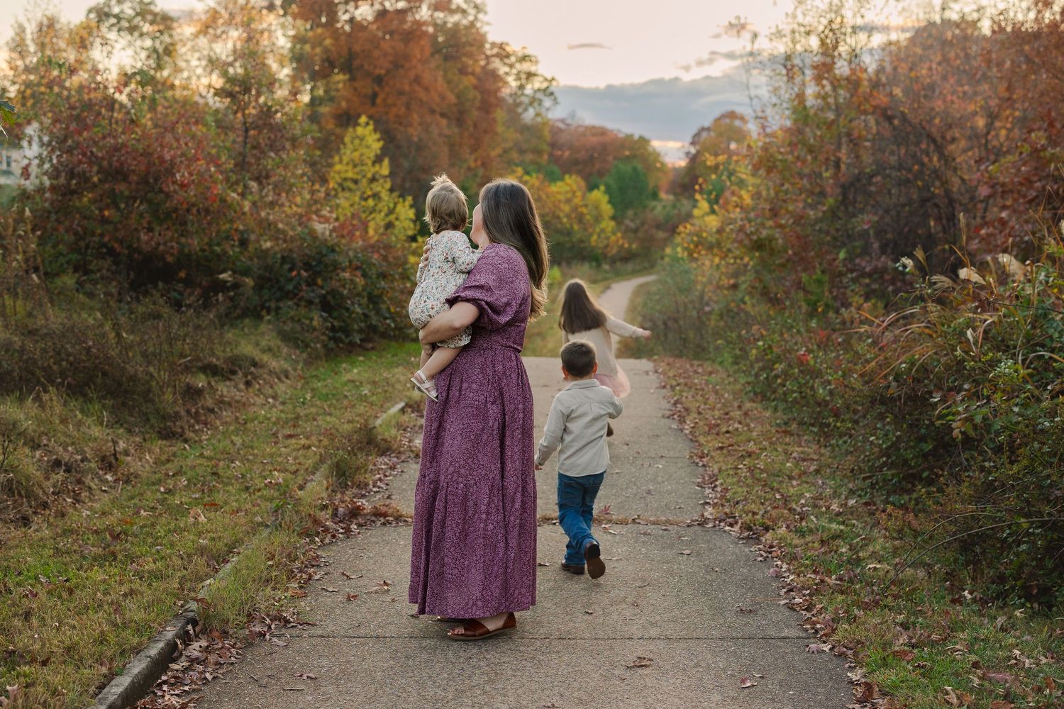 Family wearing purple dresses walks along an autumn trail surrounded by fall foliage at sunset.