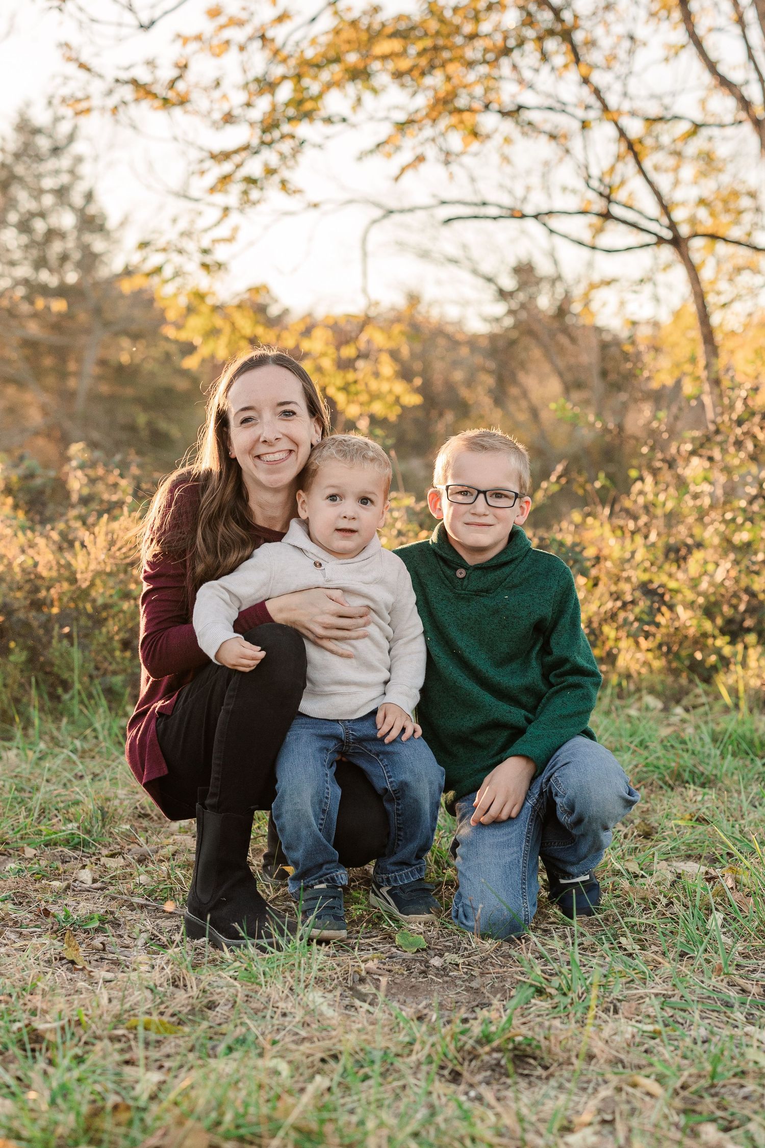 Mother and children on grassy ground during golden hour with autumn foliage in the background.
