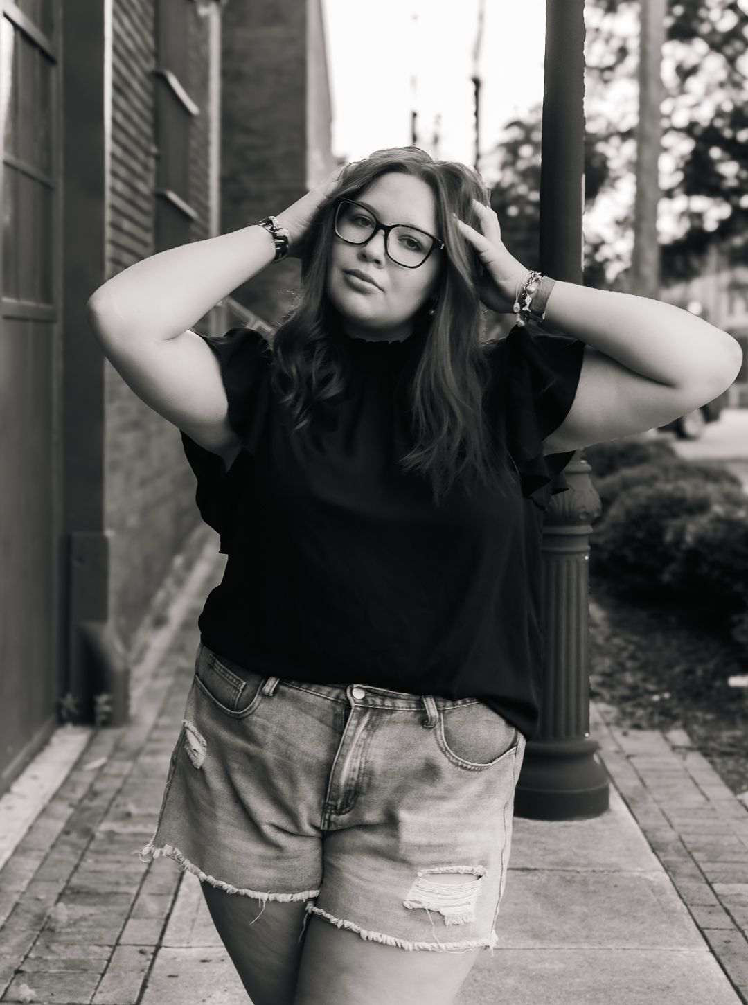 A casual black and white portrait of someone in denim shorts and glasses on a brick walkway.