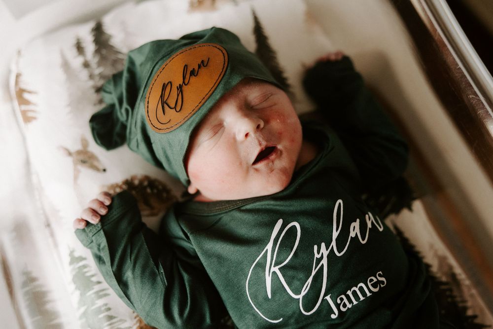 Newborn baby sleeping peacefully in a sage green swaddle blanket on a white surface.
