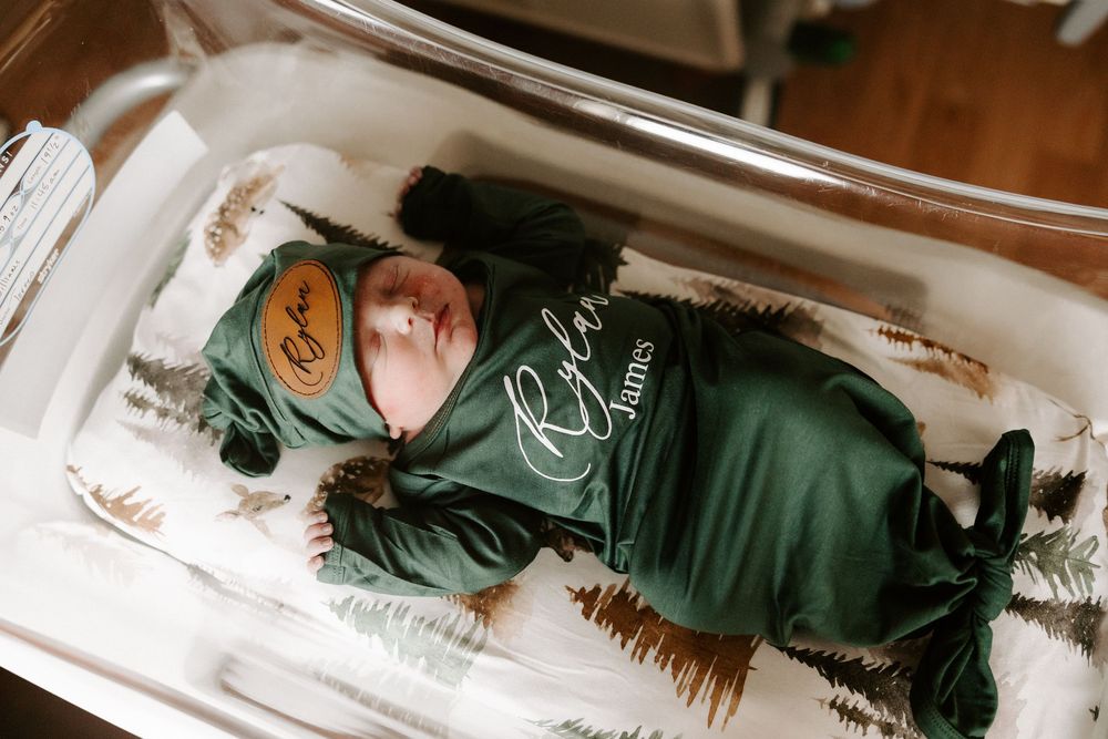 A sleeping baby wearing a dark green onesie lies on white bedding.