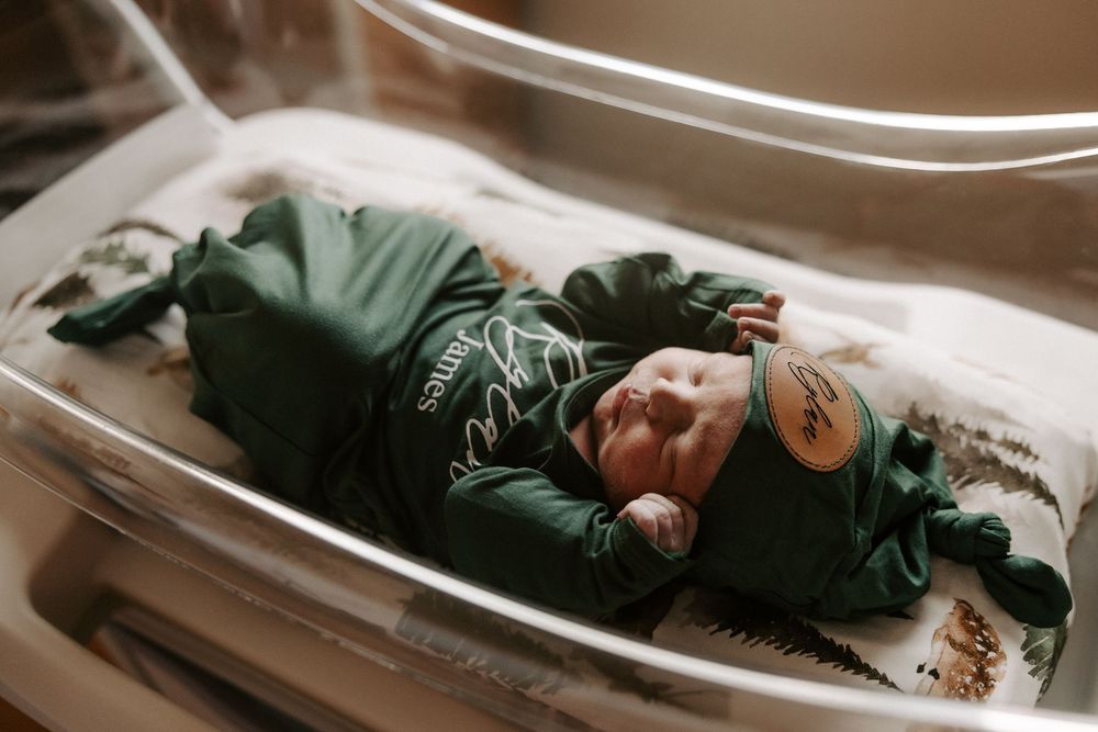 Sleeping newborn wrapped in forest green swaddle with name text overlay on hospital bassinet.