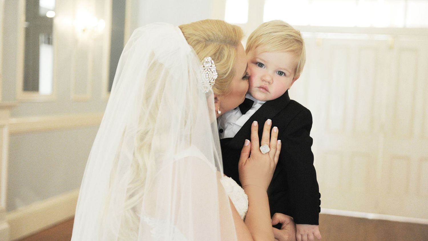 A bride in a white wedding gown and veil tenderly kisses a young blonde toddler boy in a black suit.